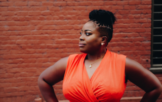 A plus-size Black woman in an orange wrap dress, with a brick wall backdrop, demonstrating a body liberation concept