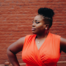 A plus-size Black woman in an orange wrap dress, with a brick wall backdrop, demonstrating a body liberation concept