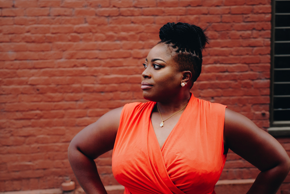A plus-size Black woman in an orange wrap dress, with a brick wall backdrop, demonstrating a body liberation concept