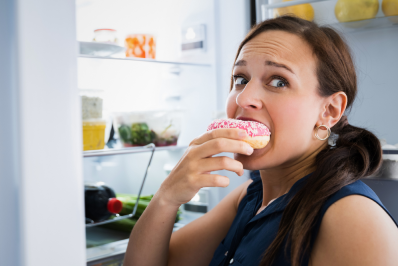 A woman engaging in emotional eating by eating a doughnut in front of an open refrigerator.