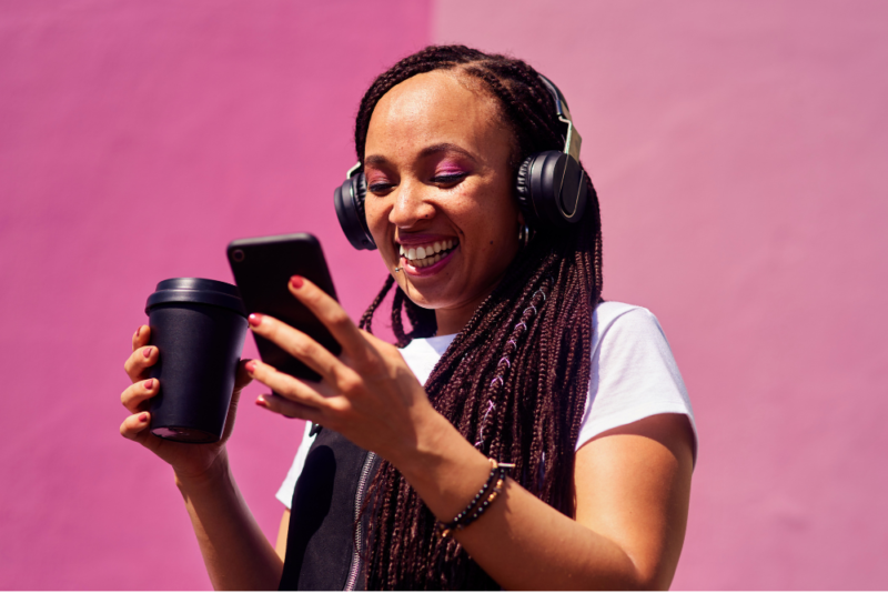 A young Black woman smiling as she drinks a cup of coffee and looks at her phone screen