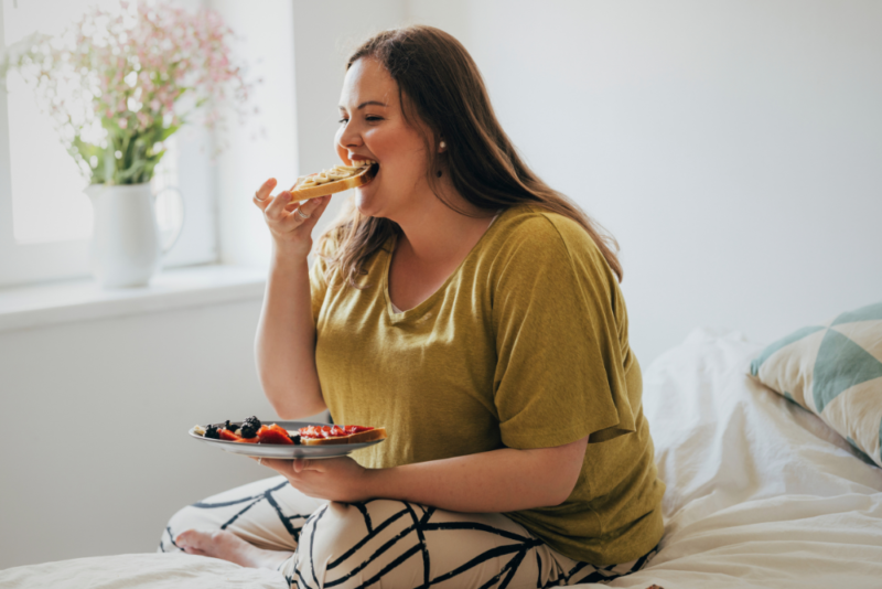 Young plus size woman sitting on her bed enjoying her breakfast.